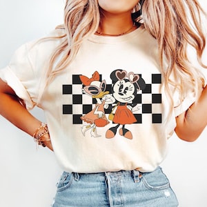 May include: Cream-colored t-shirt featuring a graphic of Minnie Mouse and Daisy Duck in front of a black and white checkered background. Minnie wears heart-shaped sunglasses and a red dress. Daisy wears a bow and a peach-colored dress.