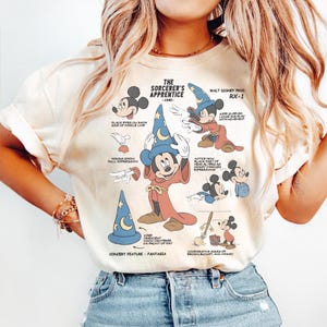 May include: Cream-colored t-shirt featuring a graphic of Mickey Mouse as the Sorcerer's Apprentice from Fantasia. The design includes illustrations and text detailing the character's features and the film's production. The text includes "The Sorcerer's Apprentice - 1940".