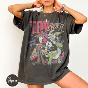 Puede incluir: Camiseta gris oscuro con un gr&aacute;fico de Toy Story. El dise&ntilde;o incluye a Woody, Buzz Lightyear y Rex, con las palabras "Toy Story" y "Since 1995". La camiseta tiene un aspecto vintage y desgastado.