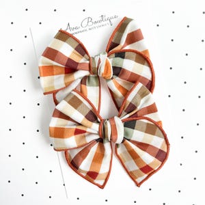 Può includere: Due fiocchi per capelli in tessuto con motivo a quadri nei toni dell'arancione, marrone, crema e verde tenue. I fiocchi sono fissati su un cartoncino bianco con la scritta Ava Bowtique Handmade Accessories stampata nella parte superiore.