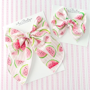Puede incluir: Dos lazos para el pelo con temática de sandía sobre tarjetas blancas. Los lazos presentan un fondo a rayas rosas y blancas con rodajas de sandía. El texto "Ava Boutique Handmade Accessories" está impreso en las tarjetas.