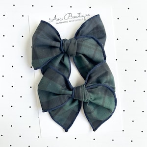 May include: Two hair bows featuring a dark green and black plaid pattern with navy blue stitched edges. The bows are attached to a white display card with the text Ava Bowtique HANDMADE ACCESSORIES printed in black at the top.