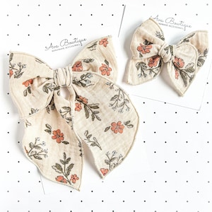 Peut inclure: Deux nœuds pour cheveux en tissu crème avec un motif floral composé de fleurs orange et de feuilles vertes. Chaque nœud est fixé sur une carte de présentation blanche avec le texte Ava Bowtique Handmade Accessories imprimé en noir.