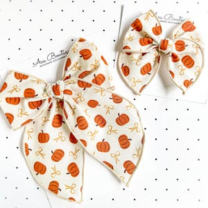 May include: Two hair bows made of off-white fabric featuring an orange pumpkin and gold ribbon print. Each bow is attached to a white card labeled Ava Bowtique Handmade Accessories. The bows have a knotted center and long, pointed tails.