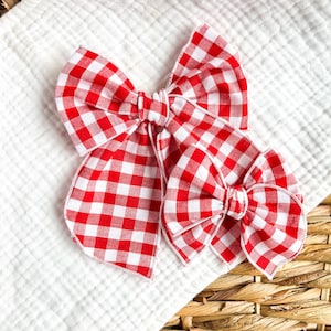 May include: Two red and white gingham hair bows. The bows are made of fabric and have a classic checkered pattern. The bows are arranged on a white, textured surface.