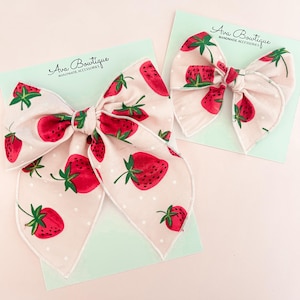 Puede incluir: Dos lazos grandes para el cabello con un estampado de fresas sobre un fondo rosa claro. Los lazos son de tela y presentan fresas rojas con hojas verdes y lunares blancos. Los lazos están sobre una tarjeta verde claro con el texto "Ava Boutique".
