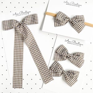 Könnte beinhalten: Eine Kollektion von vier braun-weiß karierten Haarschleifen auf weißen Karten mit der Aufschrift Ava Bowtique. Eine Schleife hat lange Bänder, eine ist an einem beigen elastischen Stirnband befestigt und zwei sind klassische Haarschleifen.