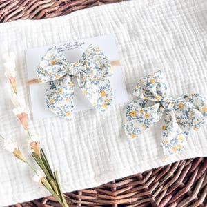 Op de afbeelding: Twee witte haarstrikken van stof met een geel bloemenpatroon. De strikken zijn gebonden met een blauw lint en bevinden zich op een witte achtergrond.
