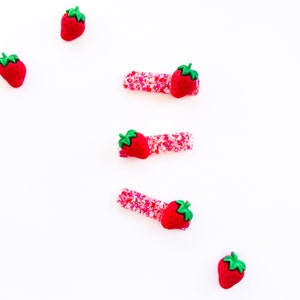 May include: A collection of hair accessories featuring a strawberry theme. The image shows several red strawberry-shaped hair clips with green stems, alongside loose strawberry charms. The clips have a pink glitter base. The items are arranged on a white background.