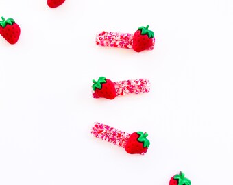 Puede incluir: Accesorios para el cabello con temática de fresas. La imagen muestra varias pinzas para el pelo rojas con forma de fresa y tallos verdes, junto con dijes de fresa sueltos. Las pinzas tienen una base de purpurina rosa. Los artículos están dispuestos sobre un fondo blanco.