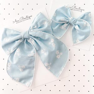 Op de afbeelding: Twee lichtblauwe stoffen haarstrikken met witte bloempatronen. De strikken zijn in het midden geknoopt en hebben lange, vloeiende uiteinden. Ze liggen op witte kaarten met de tekst "Ava Boutique HANDMADE ACCESSORIES".