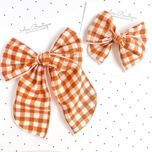 Könnte beinhalten: Zwei Haarschleifen mit einem orange-weißen Vichy-Karo-Muster. Jede Schleife ist auf einer weißen Präsentationskarte mit der Aufschrift Ava Bowtique Handmade Accessories befestigt. Die Schleifen haben eine Knotenmitte und lange Stoffbänder.
