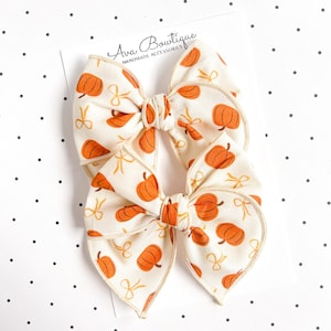 May include: Two fabric hair bows attached to a white display card labeled Ava Bowtique Handmade Accessories. The bows feature an off-white background with a repeating pattern of orange pumpkins and small yellow ribbon loops throughout the design.