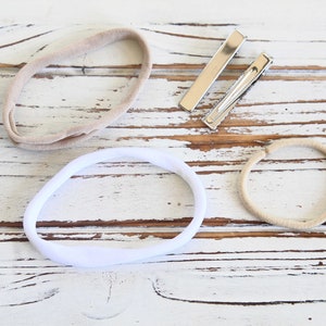 May include: Overhead view of baby hair accessories: two beige headbands, a white headband, and two silver barrette clips. The items are arranged on a distressed white wooden surface.