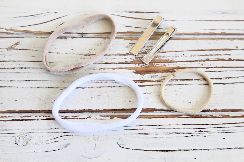 May include: Overhead view of baby hair accessories: two beige headbands, a white headband, and two silver barrette clips. The items are arranged on a distressed white wooden surface.
