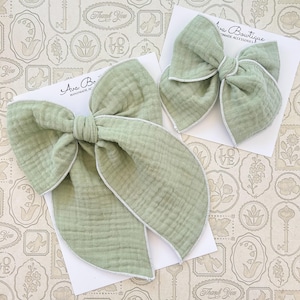 Peut inclure: Deux nœuds pour cheveux en tissu vert sauge avec bordure blanche, présentés sur des cartons blancs. Les nœuds ont un aspect texturé et superposé. Les cartes portent le texte "Ava Boutique HANDMADE ACCESSORIES". Le fond est un papier à motifs avec des timbres et les mots "Thank You" et "Love".