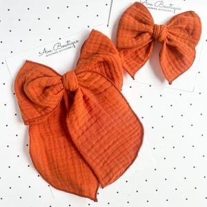May include: Two burnt orange hair bows made of textured muslin fabric, each attached to a white display card featuring the text Ava Boutique Handmade Accessories. The bows have a knotted center and long, flowing tails with visible fabric seams.
