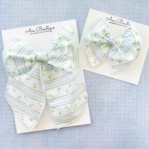 Op de afbeelding: Twee witte stoffen haarstrikken met blauwe en groene bloemen- en streepmotieven. De strikken liggen op witte kaarten met de tekst "Ava Boutique HANDMADE ACCESSORIES". Eén strik is groter dan de andere.