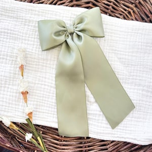 May include: A sage green satin bow with long, flowing tails. The bow is centered and tied, with the tails extending downwards. The bow is set against a white, textured fabric and a wicker basket.