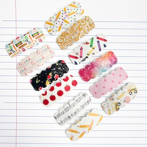 May include: A collection of twelve scalloped hair clips featuring various school-themed patterns including pencils, crayons, apples, musical notes, school buses, and glittery textures in pink, gold, and black, arranged on a lined paper background.