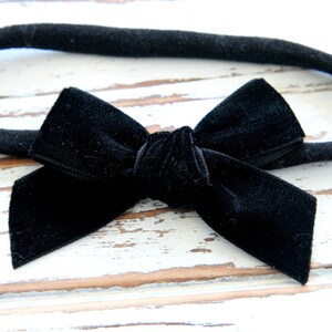 May include: A black velvet bow headband. The bow is centered on a black fabric band. The bow is a classic shape, with a knot in the center and two loops on either side. The headband is suitable for babies and children.