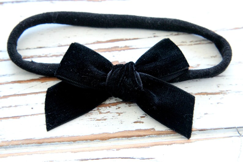 May include: A black velvet bow headband. The bow is centered on a black fabric band. The bow is a classic shape, with a knot in the center and two loops on either side. The headband is suitable for babies and children.