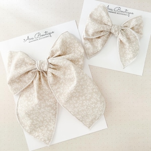 May include: Two beige fabric hair bows with a white floral pattern, displayed on white cards. The bows are tied in the center and have a contrasting white trim. The cards have the text "Ava Boutique HANDMADE ACCESSORIES".