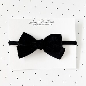 May include: A black velvet bow headband. The bow is centered on a thin black elastic band. The headband is displayed on a white card with the text "Ava Boutique HANDMADE ACCESSORIES". The background is white with small black polka dots.