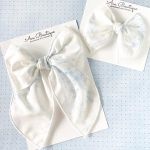 Op de afbeelding: Twee witte stoffen haarstrikken met een delicaat blauw bloemenpatroon worden gepresenteerd op witte kaarten. De strikken zijn in het midden geknoopt. De tekst "Ava Boutique HANDMADE ACCESSORIES" staat op de kaarten.