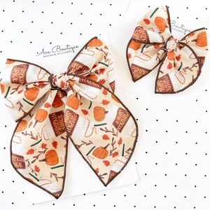 May include: Two hair bows featuring a cream-coloured fabric with a repeating pattern of orange pumpkins, brown autumn leaves, and white cups labelled Pumpkin Spice. Each bow has dark brown stitched edges and is attached to a white display card.