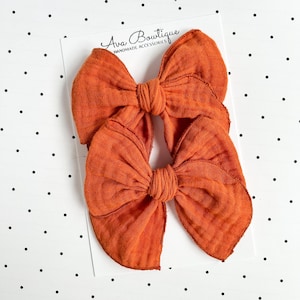 Peut inclure: Deux nœuds pour cheveux orange brûlé fabriqués dans un tissu texturé et froissé. Les nœuds sont fixés sur une carte de présentation blanche portant le texte Ava Bowtique Handmade Accessories en police noire cursive et sans-serif.