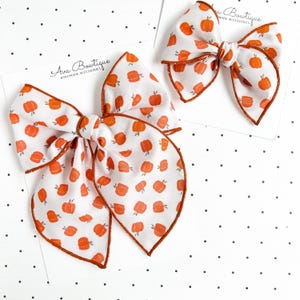Peut inclure: Deux nœuds pour cheveux en tissu blanc avec un motif de citrouilles orange et une fine bordure orange. Chaque nœud est fixé sur une carte de présentation blanche à pois noirs avec le texte Ava Bowtique Handmade Accessories en noir.