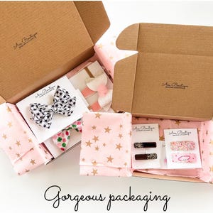 May include: Open cardboard boxes filled with hair accessories, including bows and barrettes. The items are wrapped in pink tissue paper with gold stars. One bow features a soccer ball pattern. The text "Gorgeous packaging" is at the bottom.