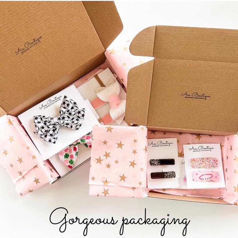May include: Open cardboard boxes filled with hair accessories, including bows and barrettes. The items are wrapped in pink tissue paper with gold stars. One bow features a soccer ball pattern. The text "Gorgeous packaging" is at the bottom.