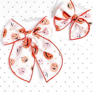 Può includere: Due fiocchi per capelli con sfondo bianco caratterizzati da un motivo ripetuto di zucche di Halloween arancioni e rosa chiaro e piccole stelle. Ogni fiocco ha un bordo profilato in arancione brillante ed è fissato su un cartoncino con l'etichetta Ava Bowtique Handmade Accessories.
