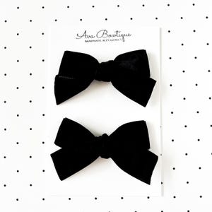 May include: Two black velvet hair bows on a white card with the text "Ava Boutique HANDMADE ACCESSORIES". The bows are tied in a classic bow shape, perfect for adding a touch of elegance to any hairstyle.