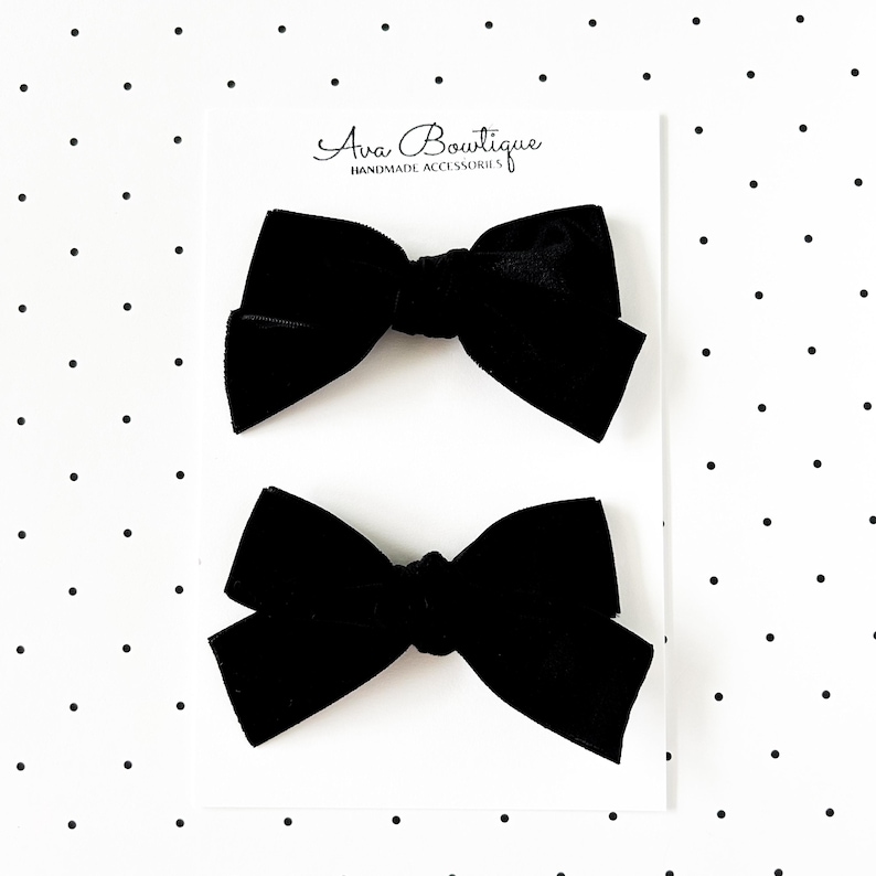 May include: Two black velvet hair bows on a white card with the text "Ava Boutique HANDMADE ACCESSORIES". The bows are tied in a classic bow shape, perfect for adding a touch of elegance to any hairstyle.