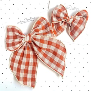 May include: Two hair bows featuring a burnt orange and cream gingham check pattern. Each bow is attached to a white display card with black polka dots and the text Ava Bowtique Handmade Accessories. The bows have frayed edges and a center knot.