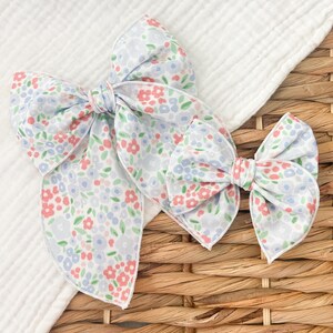 Op de afbeelding: Drie stoffen haarstrikken met een bloemenpatroon. De strikken tonen kleine rode, blauwe en groene bloemen op een witte achtergrond. De strikken zijn gerangschikt op een witte doek en een rieten mand.