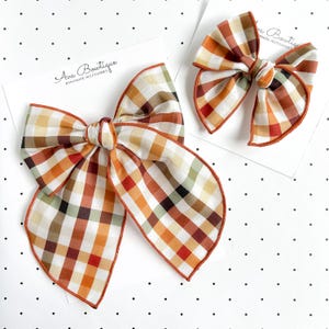 Könnte beinhalten: Zwei Haarschleifen mit Karomuster in Orange-, Braun-, Creme- und Grüntönen. Jede Schleife ist auf einer weißen Karte befestigt, auf der der Text Ava Bowtique Handmade Accessories in Schwarz gedruckt ist. Die Schleifen haben einen rostfarbenen Rand.