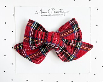 May include: A red plaid hair bow with black, white, and yellow stripes, attached to a white display card featuring the text Ava Bowtique HANDMADE ACCESSORIES. The fabric bow has a knotted center and pointed ends, showcasing a classic tartan pattern.