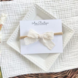 A cream-colored baby headband with a delicate lace bow. The bow is attached to a tan elastic band