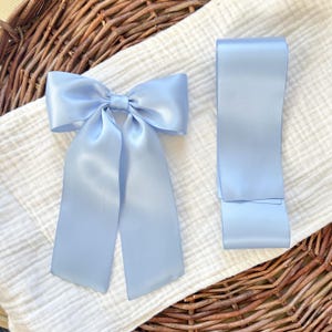 May include: A light blue satin bow with long streamers and a matching ribbon. The bow is tied and sits on a white, textured fabric. The ribbon is folded and lies next to the bow. The background is a woven basket.