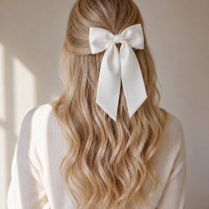 Könnte beinhalten: Ein großer, weißer Satin-Haarclip mit Schleife ist im Haar befestigt. Die Schleife ist im klassischen Stil mit langen, fließenden Enden gebunden. Das Haar ist lang, wellig und blond. Die Person trägt einen cremefarbenen Pullover.