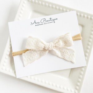 May include: A cream lace bow headband with a tan elastic band, presented on a white card with the text "Ava Boutique HANDMADE ACCESSORIES". The bow is on a white square plate.