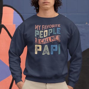May include: Navy blue sweatshirt with the text "MY FAVORITE PEOPLE CALL ME PAPI" in a retro, multi-colored design. The sweatshirt is a solid color and has a crew neck.