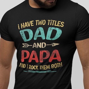 Pode incluir: Camiseta preta com o texto "I HAVE TWO TITLES DAD AND PAPA AND I ROCK THEM BOTH" em verde-azulado, vermelho e amarelo. A camiseta tem um visual desgastado e vintage, com um gráfico de seta.