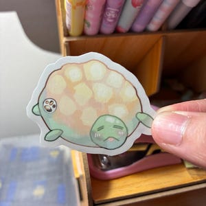 May include: A die-cut sticker featuring a cartoon turtle with a sad expression. The turtle has a light green body, a yellow and peach shell, and small green flippers. The sticker is held in front of a wooden organizer with pens.