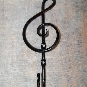 May include: Black metal treble clef wall hook. The hook is shaped like a musical treble clef symbol. The hook is made of wrought iron and has a curved hook at the bottom. The hook is designed to hang items.