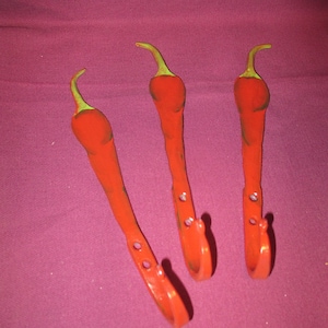 May include: Three red chili pepper-shaped metal hooks with green stems. The hooks are mounted on a burgundy background. Each hook has a curved bottom and two holes for mounting.